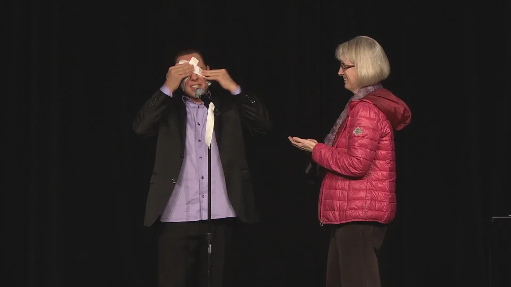 Randy wiping face as part of an act on stage with a woman standing beside him.