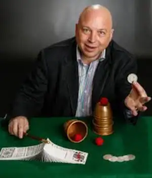 Clive The Conjuror, holding a coin while sitting, with some cards placed in front of him on the desk.